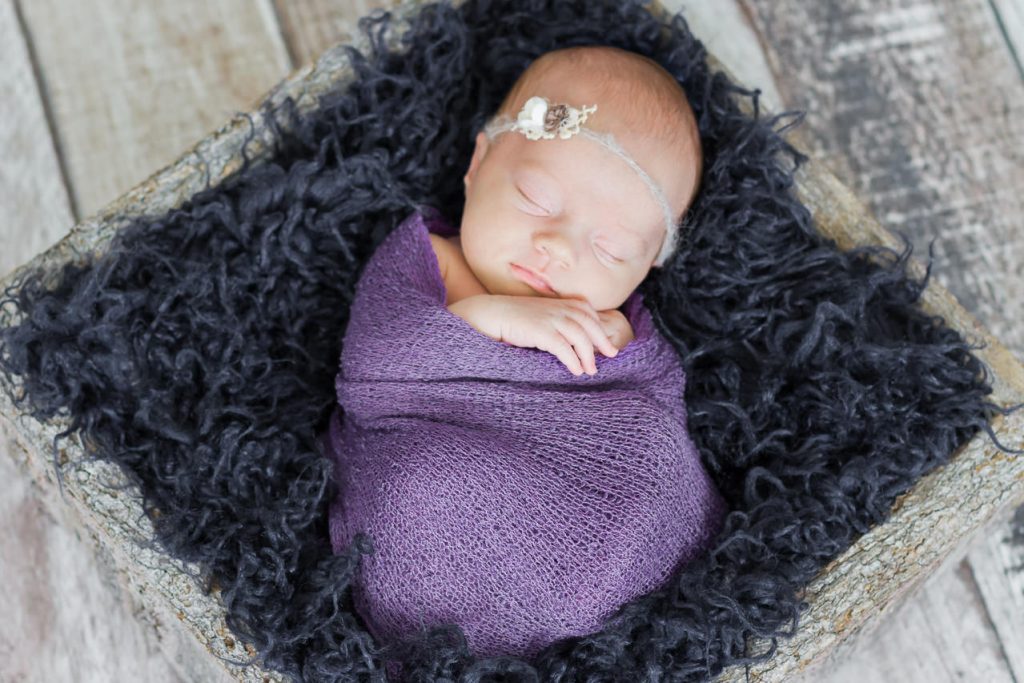 Neugeborenes liegt auf dunkel blauem fell und schläft. es wurde in ein lilafarbenes tuch gepuckt . Neugeborenenfotografie ella photo düsseldorf
