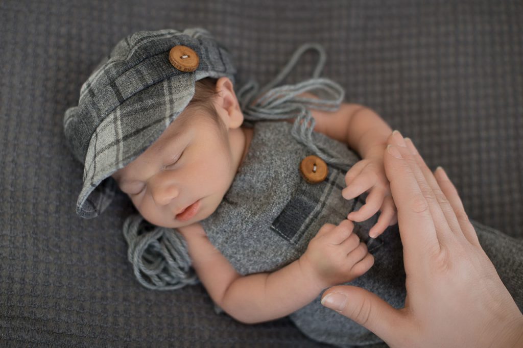 Neugeborenes liegt auf einer grauen decke und schläft. es trägt eine graue cappi und einen karierten einteiler. Neugeborenenfotografie ella photo düsseldorf