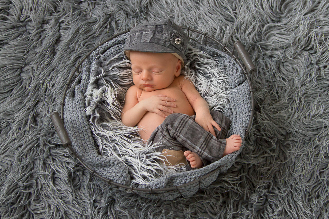 Neugeborenes liegt auf grauen fell und schläft. es trägt eine graue cappi und eine karierte Hose. Neugeborenenfotografie ella photo düsseldorf