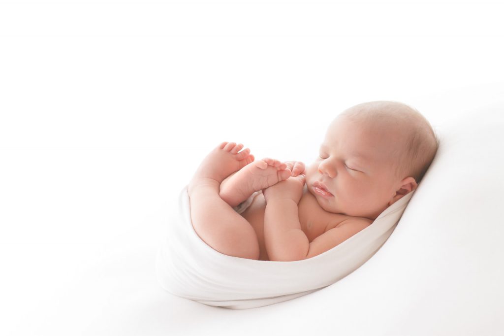 neugeborenes liegt in einen weißes tuch auf einer weissen decke und schläft, baby fotos ella photography mettmann