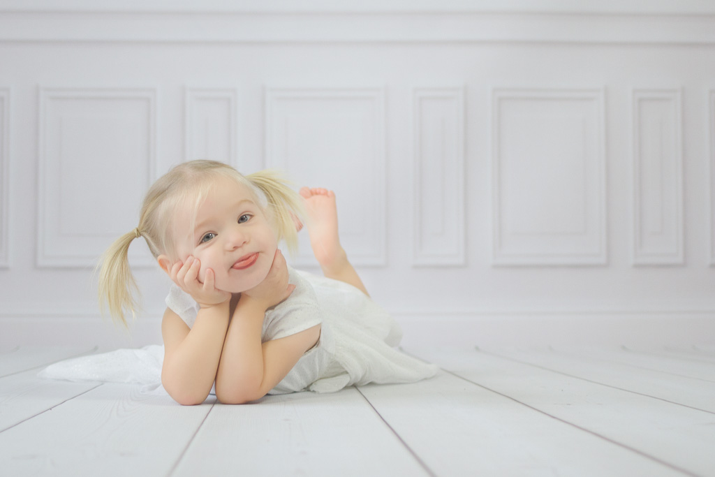 Kleines Mädchen liegt auf einem weißen Holzboden und streckt die Zunge raus. Kinderfotos Erkrath von Ella Faust