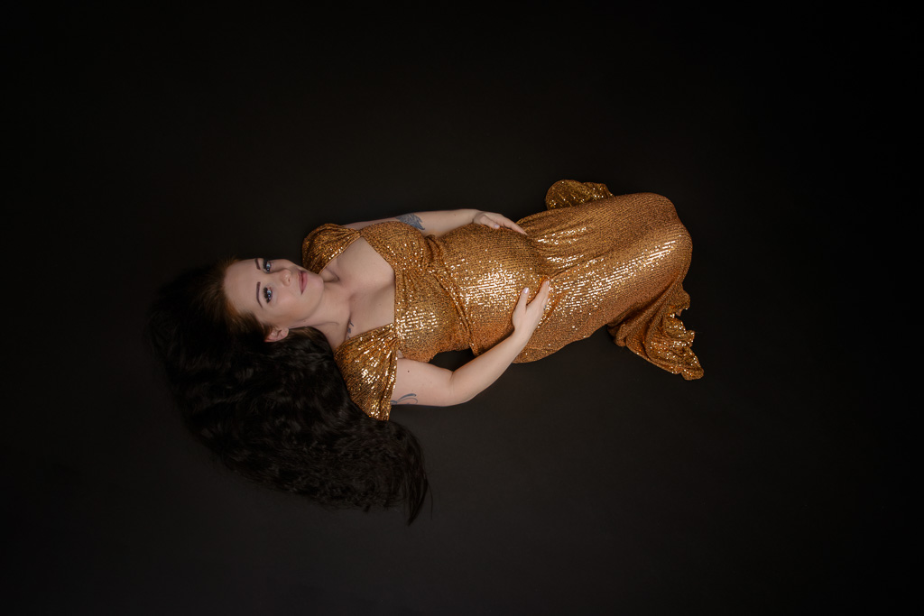 Schwangere in einen goldenen Paillettenkleid. sie liegt auf den Boden und schaut zur Kamera, Schwangerschaftsfotos von Ella Faust