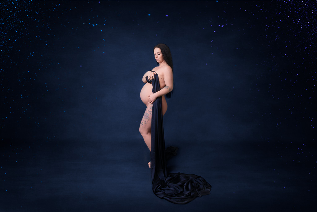 Schwangere steht mit blauem Seidenschal vor blauem Hintergrund mit Glitzer in den Ecken. Schwangerschaft Akt von Ella Faust. Ella Photography bei Düsseldorf.
