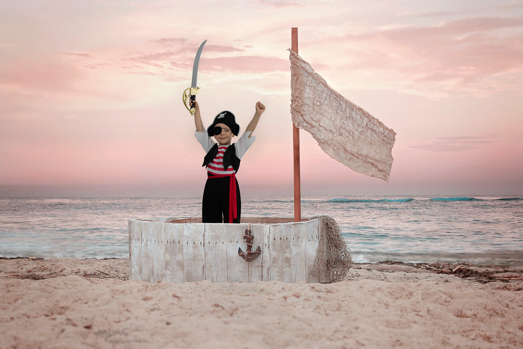 ein junge steht in einem piratenschiff und nimmt die hände hoch, das schiff steht am strand. kinderfotos ella photography
