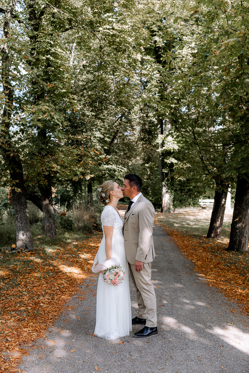 Brautpaar steht auf einer Baumallee und küsst sich